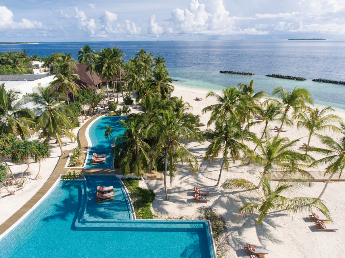 Ansicht von oben Ein tropisches Resort mit einem großen, geschwungenen Pool umgeben von Palmen, angrenzend an einen weißen Sandstrand mit Blick auf einen ruhigen, blauen Ozean. Sonnenliegen stehen sowohl am Pool als auch am Strand bereit.