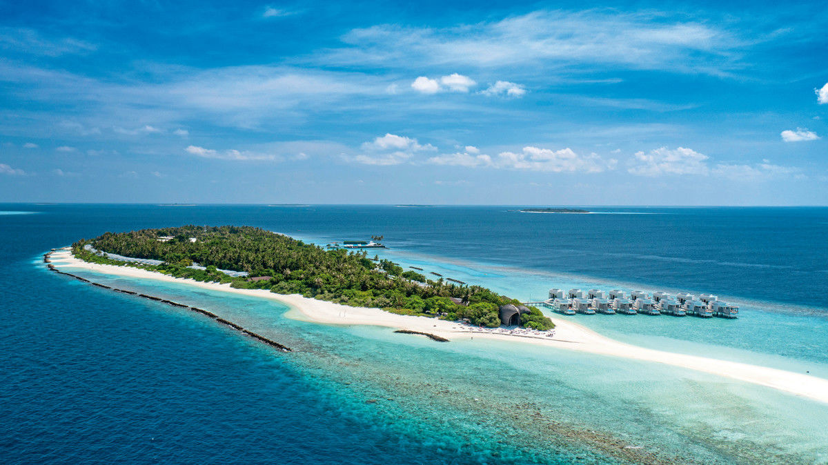 Außenansicht Luftaufnahme einer tropischen Insel mit üppiger Vegetation und einem Resort mit Überwasser-Bungalows. Türkisfarbenes Wasser umgibt die Insel, weiße Sandstrände sind erkennbar und einige Wolken liegen am Himmel.