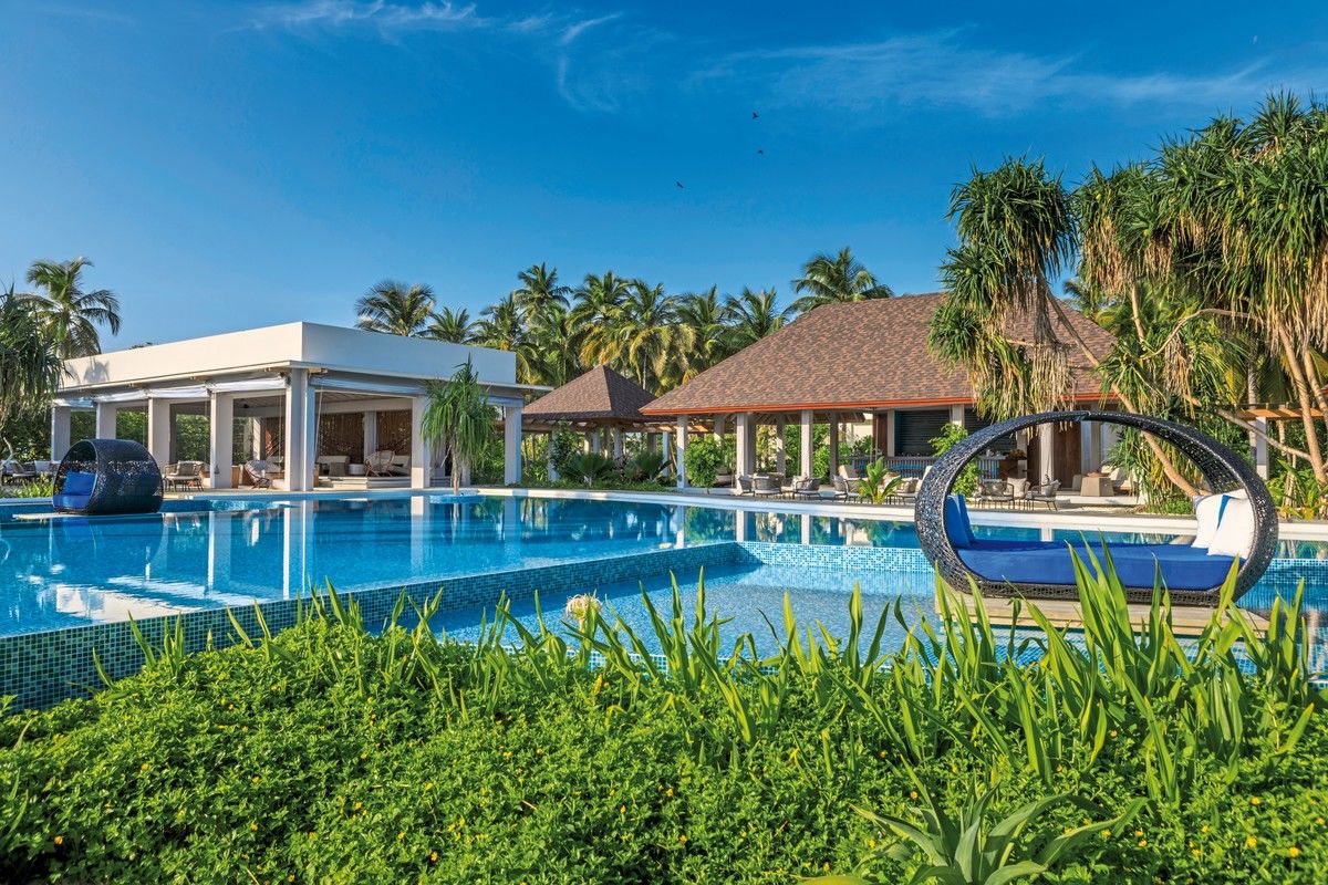 Pool Ein luxuriöses Resort mit einem großen Schwimmbad, umgeben von üppigen Grünflächen und Palmen. Im Vordergrund befindet sich eine moderne Sitzschaukel über dem Wasser, während im Hintergrund stilvolle Villen mit Ziegeldächern zu sehen sind. Der Himmel ist klar und blau, was auf ideales Urlaubswetter hinweist.