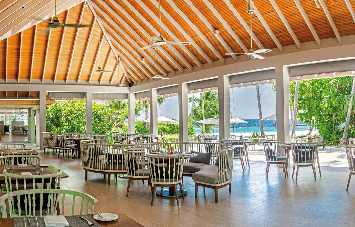 Restaurant Alt-Text: Ein helles und einladendes Strandrestaurant mit modernen Stühlen und Tischen, Holzdecken und deckenhohen Fenstern, die einen spektakulären Blick auf weißen Sandstrand, Palmen und das türkisblaue Meer freigeben.