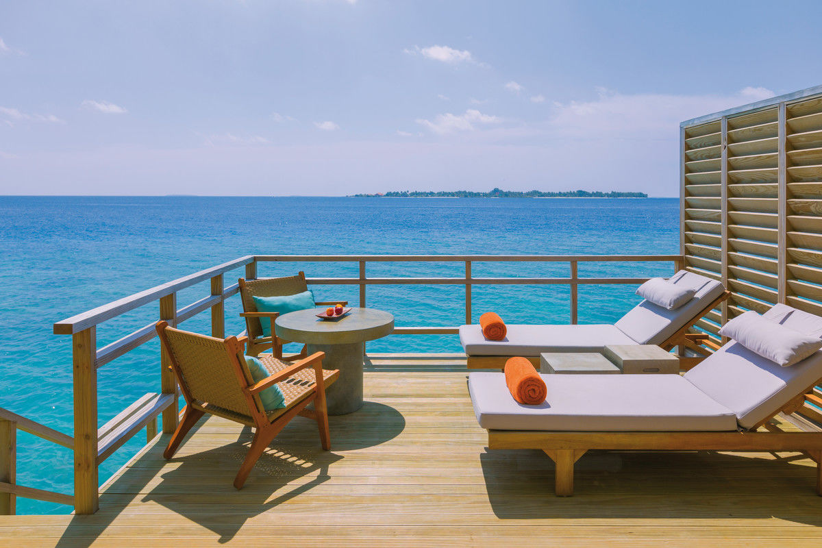 Terrasse Alt-Text: Eine entspannende Terrasse über dem Meer mit zwei Sonnenliegen, einem Tisch und Stühlen. Das kristallblaue Wasser und die Aussicht auf eine ferne Inselgruppe vermitteln ein Gefühl von Ruhe und Exklusivität. Die Handtücher auf den Sonnenliegen fügen eine einladende Note hinzu, während die Sonne für eine helle und warme Atmosphäre sorgt.