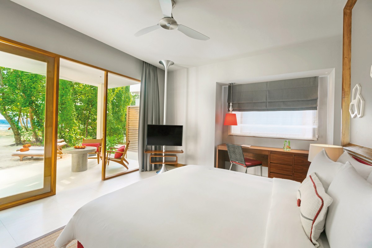 Zimmer Helles und einladendes Schlafzimmer in einem Strandresort mit einem großen Bett im Vordergrund und einer offenen Terrassentür, die den Blick auf grüne Pflanzen und den Strand freigibt. Ein Deckenventilator, Flachbildfernseher und Arbeitsbereich vervollständigen das moderne Zimmer.