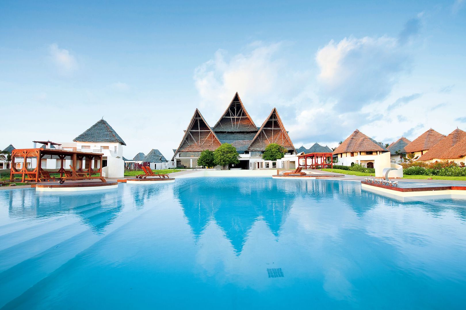 Ein luxuriöser Resort-Pool mit klarem blauen Wasser, umgeben von eleganten Liegestühlen und einer Bar im Vordergrund, während im Hintergrund traditioneller Architektur mit strohgedeckten Dächern unter einem strahlend blauen Himmel zu sehen ist.