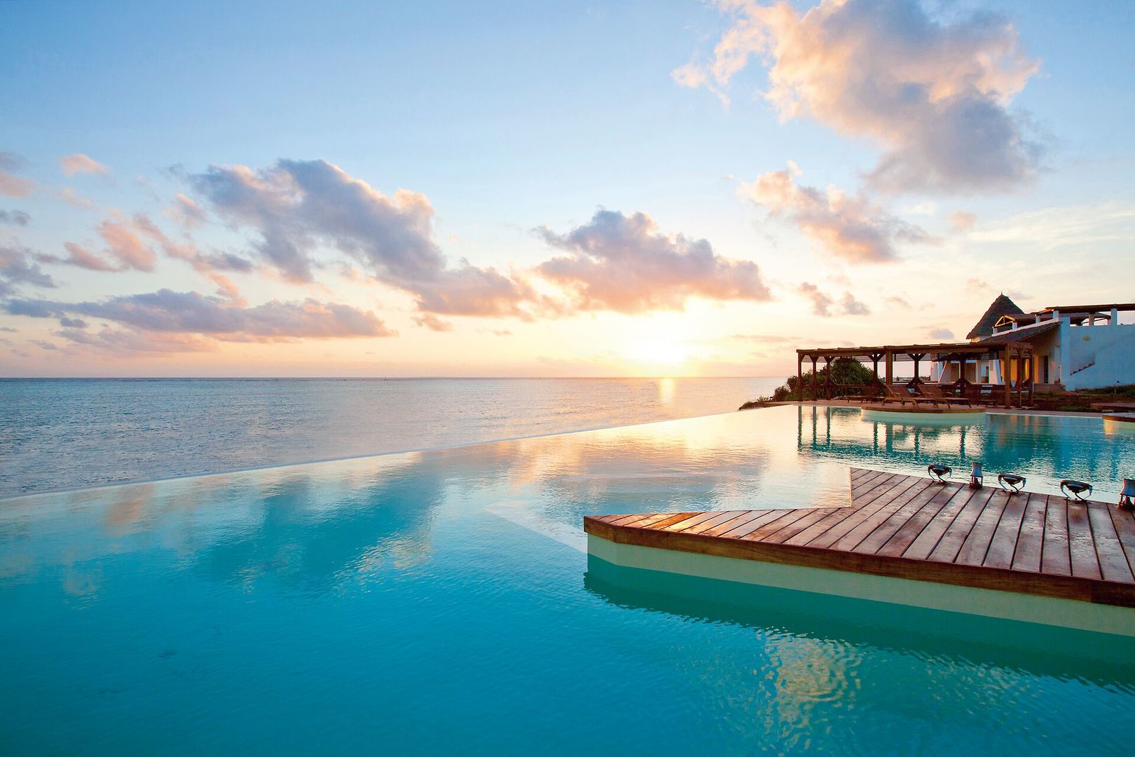 Ein luxuriöser Infinity-Pool, der nahtlos in den Ozean überzugehen scheint, unter dem farbenprächtigen Himmel während des Sonnenuntergangs. Am rechten Bildrand steht eine stilvolle Villa mit einer überdachten Terrasse, die das warme Licht der untergehenden Sonne einfängt.