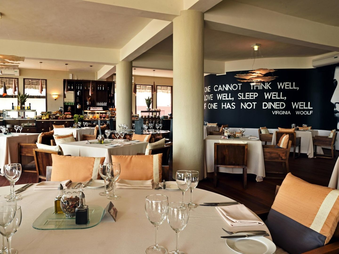 Ein elegantes Restaurantinterieur mit gedeckten Tischen, Weingläsern und bequemen Stühlen. Die Wände sind dekorativ gestaltet, insbesondere mit einem großen Zitat von Virginia Woolf: "One cannot think well, love well, sleep well, if one has not dined well." Einladende Atmosphäre ohne sichtbare Personen.
