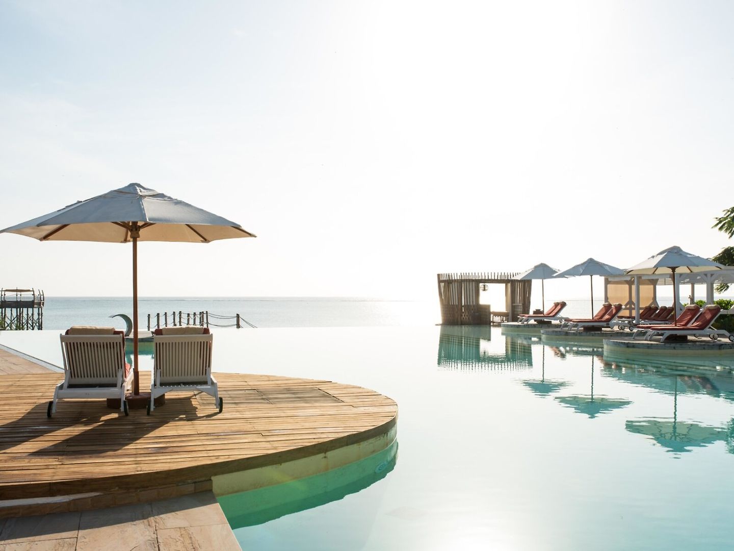 Ein luxuriöses Infinity-Pool mit Blick auf das Meer bei Tageslicht. Im Vordergrund stehen zwei Sonnenliegen unter einem geöffneten Sonnenschirm auf einem Holzdeck. Weitere Liegestühle und Sonnenschirme sind im Hintergrund entlang des Pools aufgestellt, der nahtlos in den Horizont überzugehen scheint.