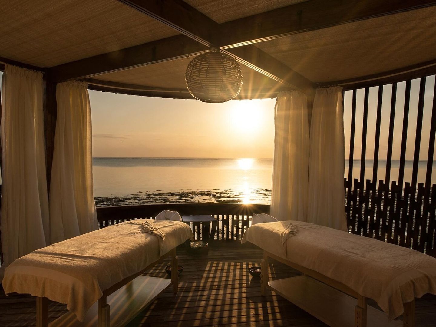 Blick auf einen entspannenden Spa-Bereich am Strand während des Sonnenuntergangs mit zwei Massagebetten, weißen Vorhängen und einem ruhigen Meer im Hintergrund.
