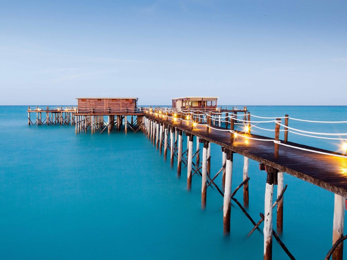Ein langer Holzsteg mit Geländern, die mit leuchtenden Lichterketten dekoriert sind, führt über das ruhige, blau schimmernde Meer zu einem Pavillon am Ende des Stegs während der Dämmerung.