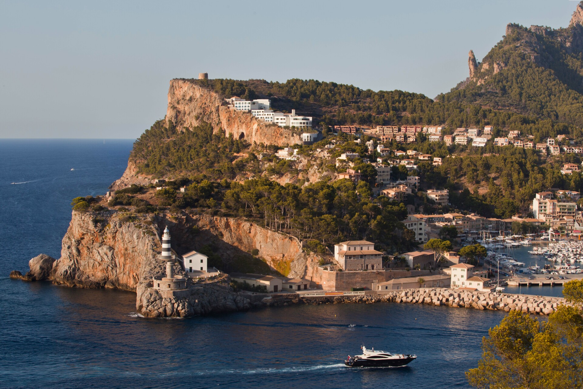 Blick auf das Jumeirah Mallorca Ein Panoramablick auf eine malerische Küstenlandschaft mit einem Leuchtturm am Vorsprung einer Klippe, einem dahinterliegenden Hügel mit dichter Bebauung und einem Yachthafen. Im Vordergrund ist eine private Yacht zu sehen, die entlang des klaren, blauen Wassers fährt.
