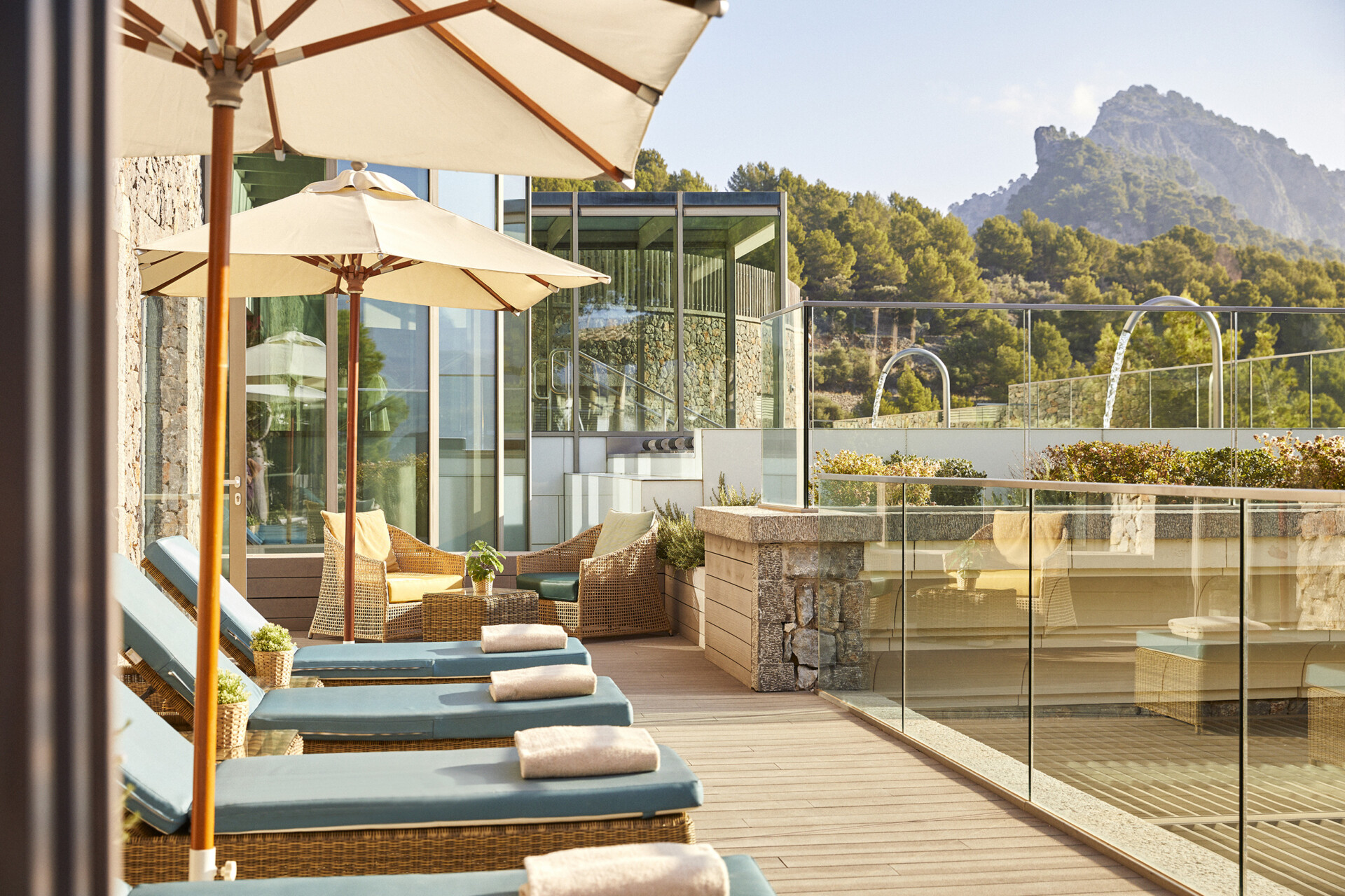 Einladende Terrasse eines Luxushotels mit Sonnenliegen unter Sonnenschirmen, gemütlichen Sitzgelegenheiten und einem Blick auf grüne Berge im Hintergrund.