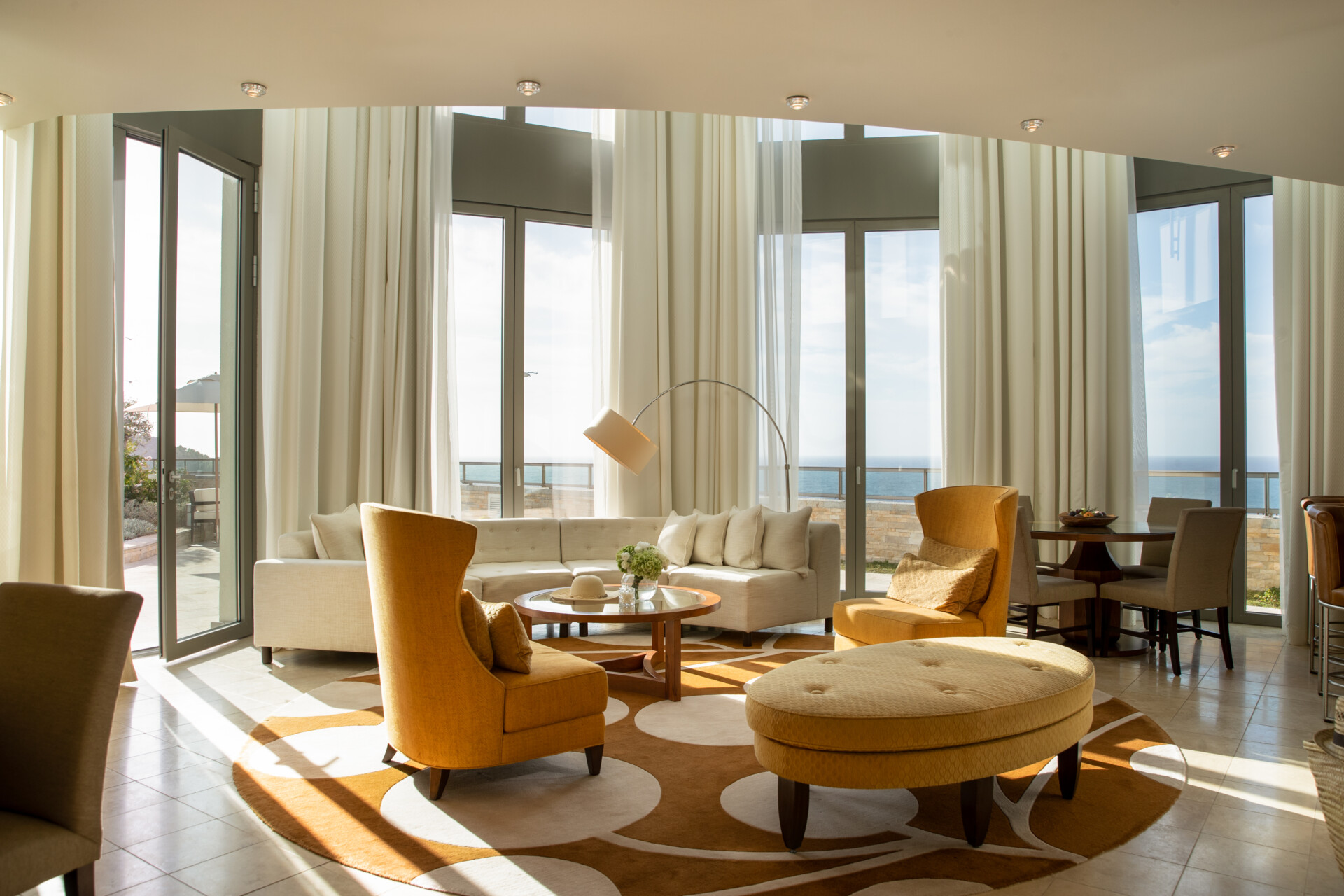 Eine geräumige und helle Lounge eines Luxushotels mit bodentiefen Fenstern und einem atemberaubenden Blick auf das Meer. Die Inneneinrichtung umfasst elegante, komfortable Möbel in warmen Erdfarben, einen großen runden Couchtisch im Zentrum, flankiert von einem beige Sofa und zwei gelben Sesseln. Weiße Vorhänge komplementieren den Raum, während eine moderne Stehlampe mit Bogen für zusätzliches Licht sorgt.