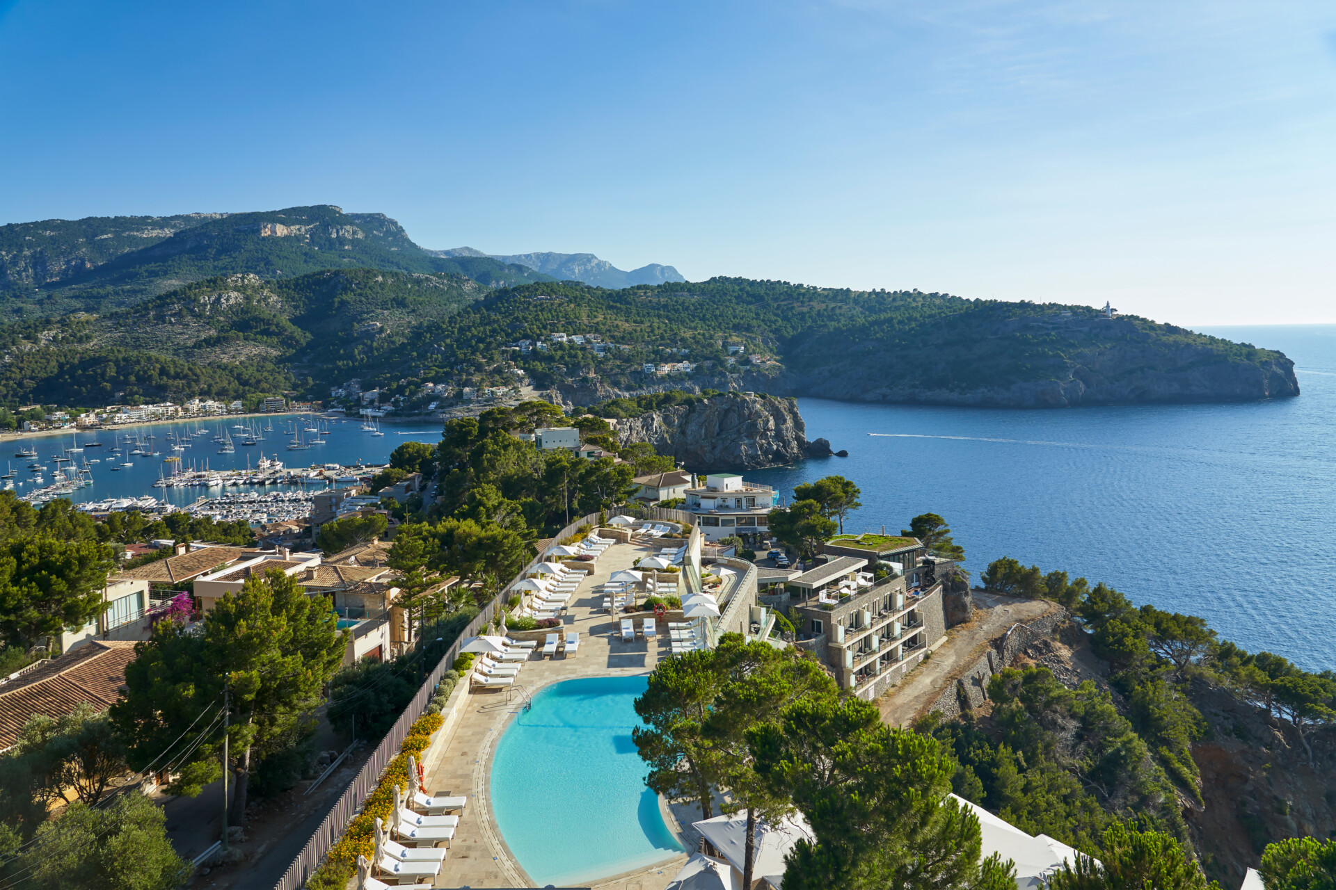 Pool mit Sonnenterrasse Blick auf eine malerische Bucht mit einem Jachthafen, umgeben von bewaldeten Hügeln und Häusern, im Vordergrund ein luxuriöses Hotel mit Pool und Liegestühlen unter strahlend blauem Himmel.