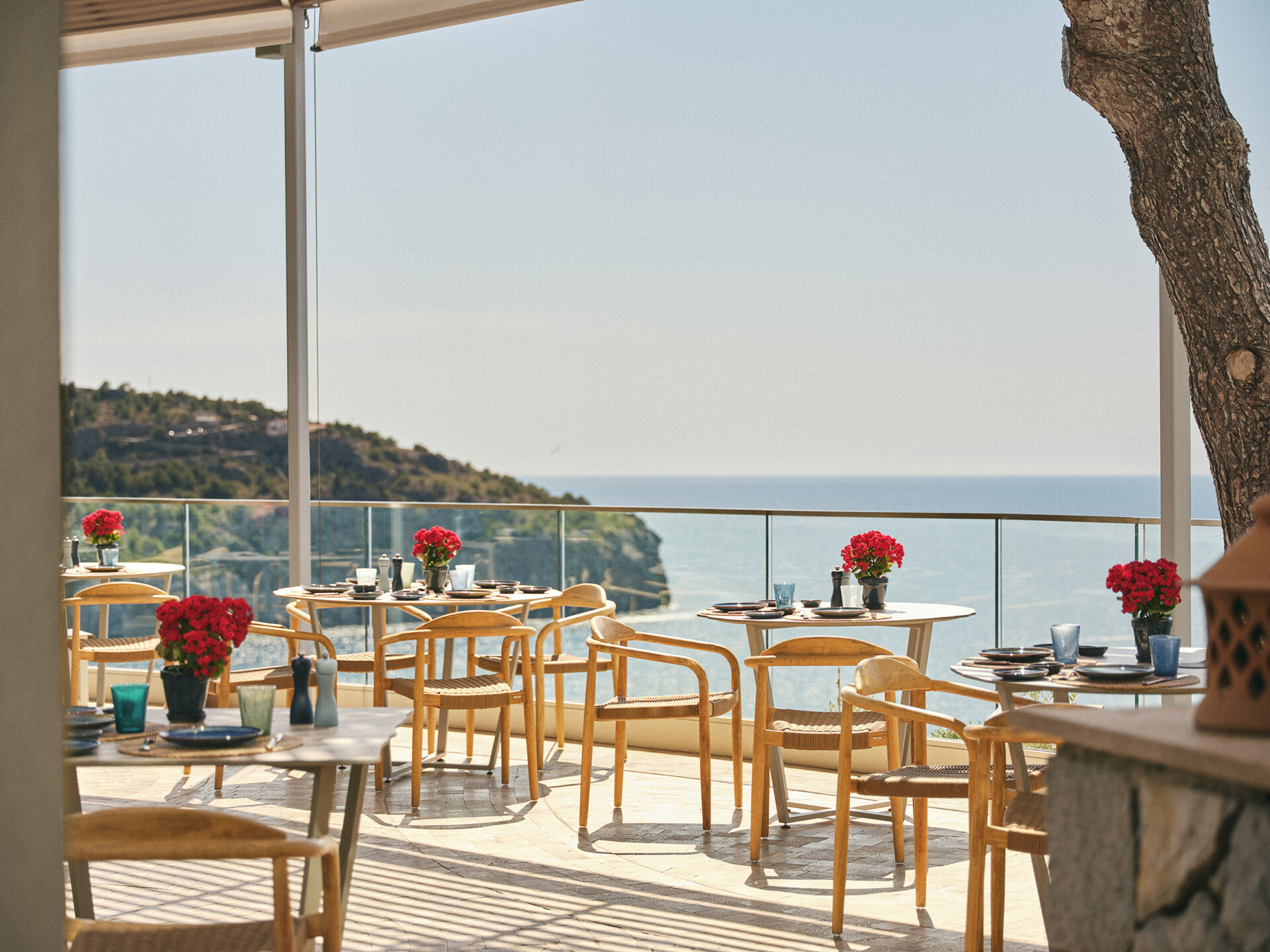 Außenterrasse eines Cafés mit Blick auf das Meer, ausgestattet mit runden Tischen, Holzstühlen und roten Blumen als Tischdeko. Klares Wetter sorgt für eine sonnige und einladende Atmosphäre.