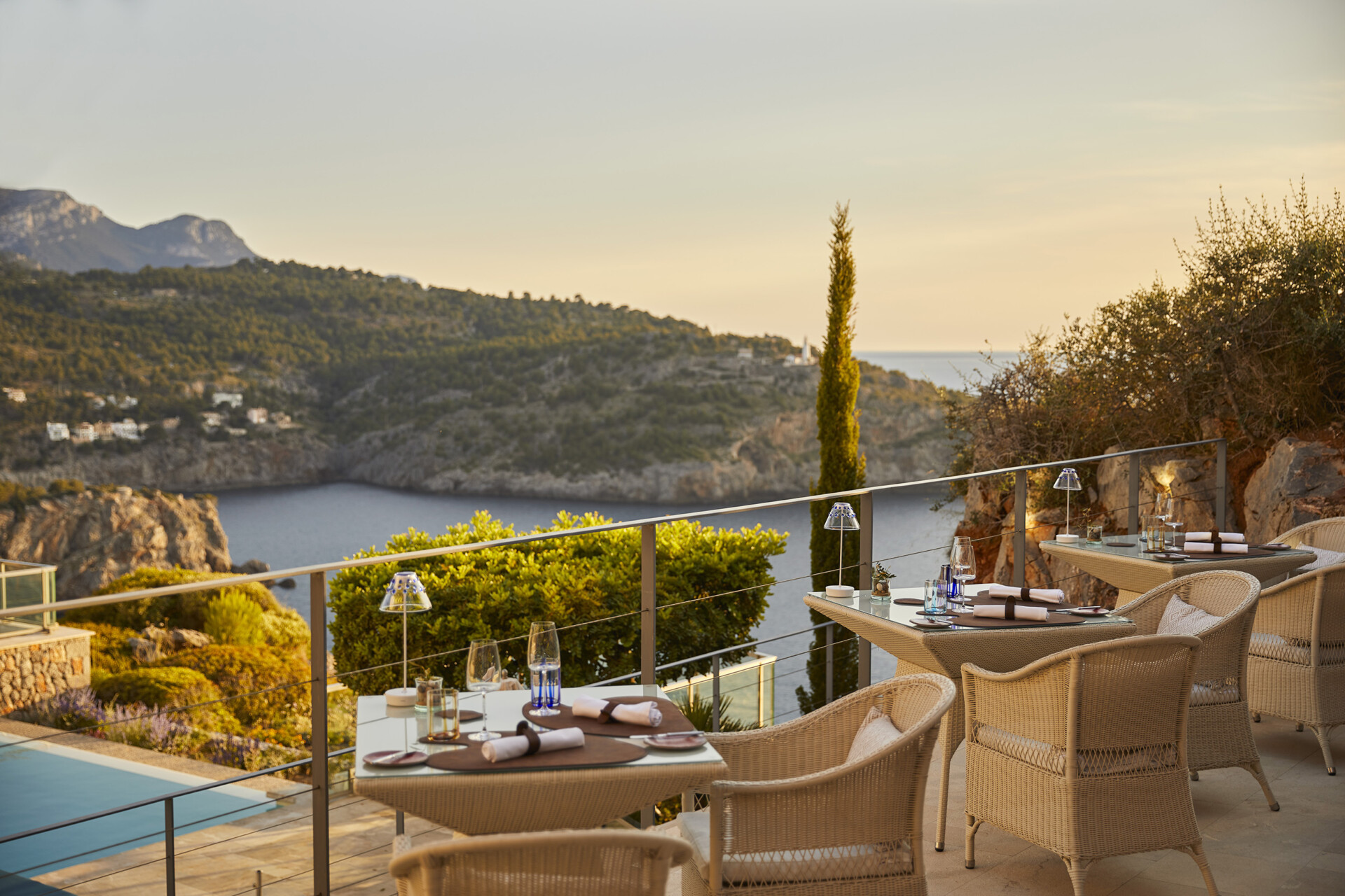 Restaurant Es Fanals Eine Außenterrasse eines Restaurants mit elegant gedeckten Tischen und Rattanstühlen. Die Terrasse bietet einen atemberaubenden Blick auf eine felsige Küstenlandschaft mit mediterraner Vegetation und das Meer bei Sonnenuntergang.