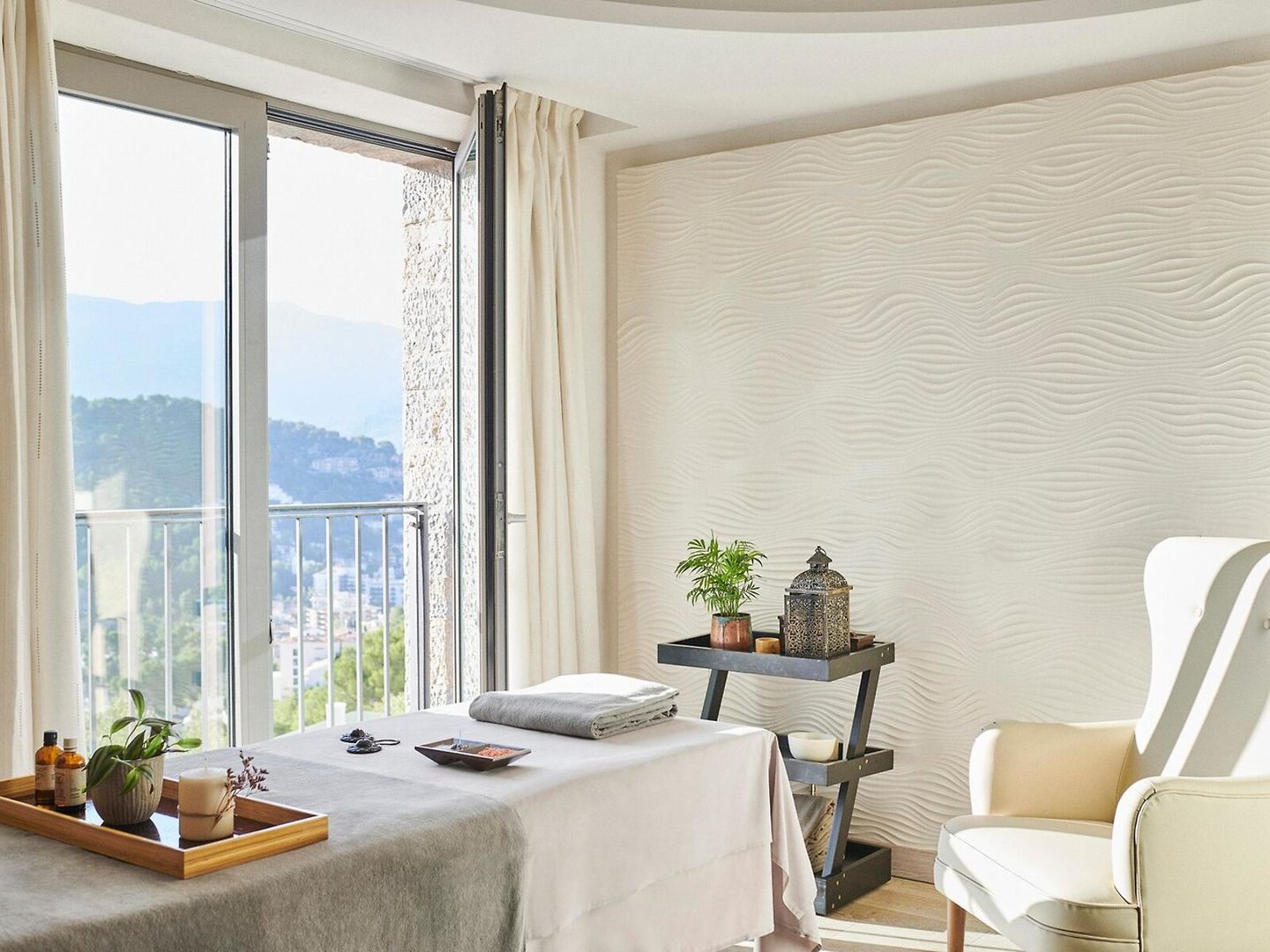 Ein hell erleuchtetes, stilvolles Zimmer mit großen Fenstern, die einen Blick auf eine hügelige Landschaft ermöglichen. Eine Massageliege ist im Vordergrund mit Handtüchern und Pflegeprodukten auf einem Holztablett zu sehen. Rechts steht ein weißer Sessel neben einem schmalen Regal mit Pflanzen und Dekorationsgegenständen. Die Wände zeigen ein modernes Wellenmuster und vermitteln eine ruhige Atmosphäre.