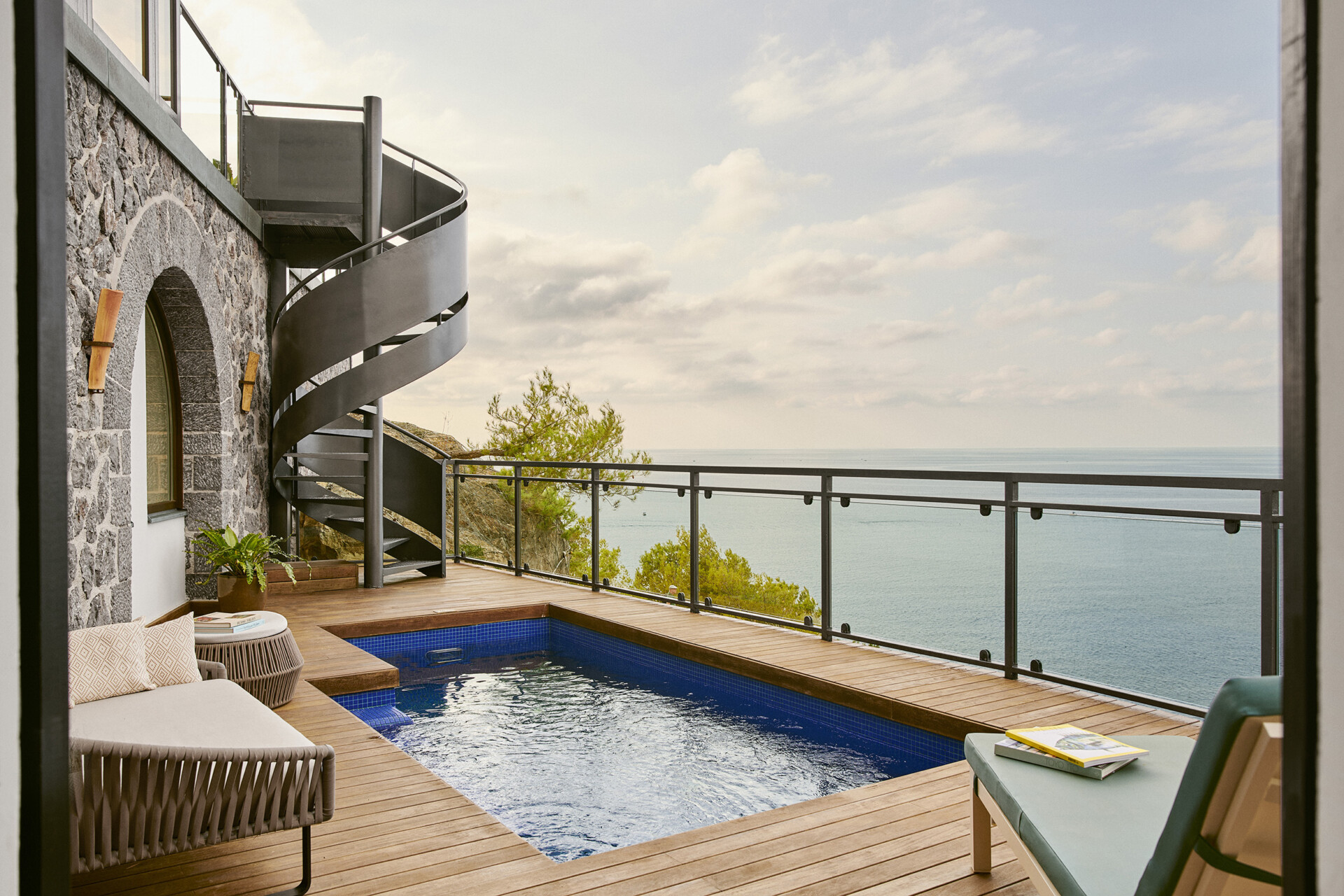 Ein eleganter Außenbereich eines Hauses mit blick auf das Meer. Im Vordergrund eine Holzterrasse mit einem kleinen Pool, daneben eine gemütliche Liege und ein runder Beistelltisch. An der Steinfassade des Hauses führt eine moderne Wendeltreppe nach oben. Im Hintergrund das ruhige Meer und ein bewölkter Himmel, eingefasst von einem schlichten Metallgeländer, das einen ungehinderten Blick auf die Natur bietet.