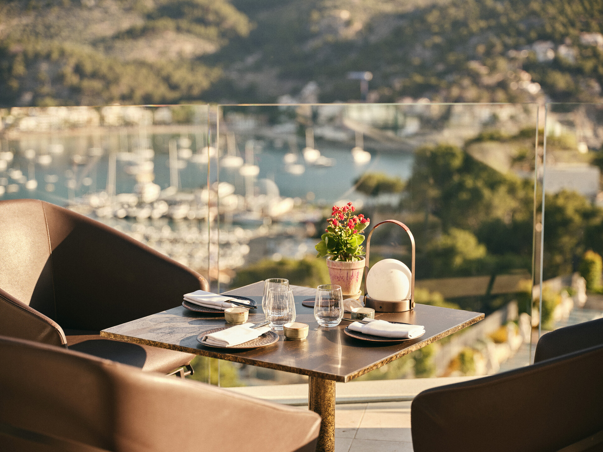 Sunset Lounge Ein gedeckter Tisch für zwei Personen auf einer Außenterrasse mit Blick auf einen malerischen Jachthafen und grüne Hügel im Hintergrund. Auf dem Tisch steht ein Blumentopf mit roten Blumen, und die Szene wird von einem warmen, sonnigen Licht beleuchtet, welches eine einladende und entspannte Atmosphäre schafft.