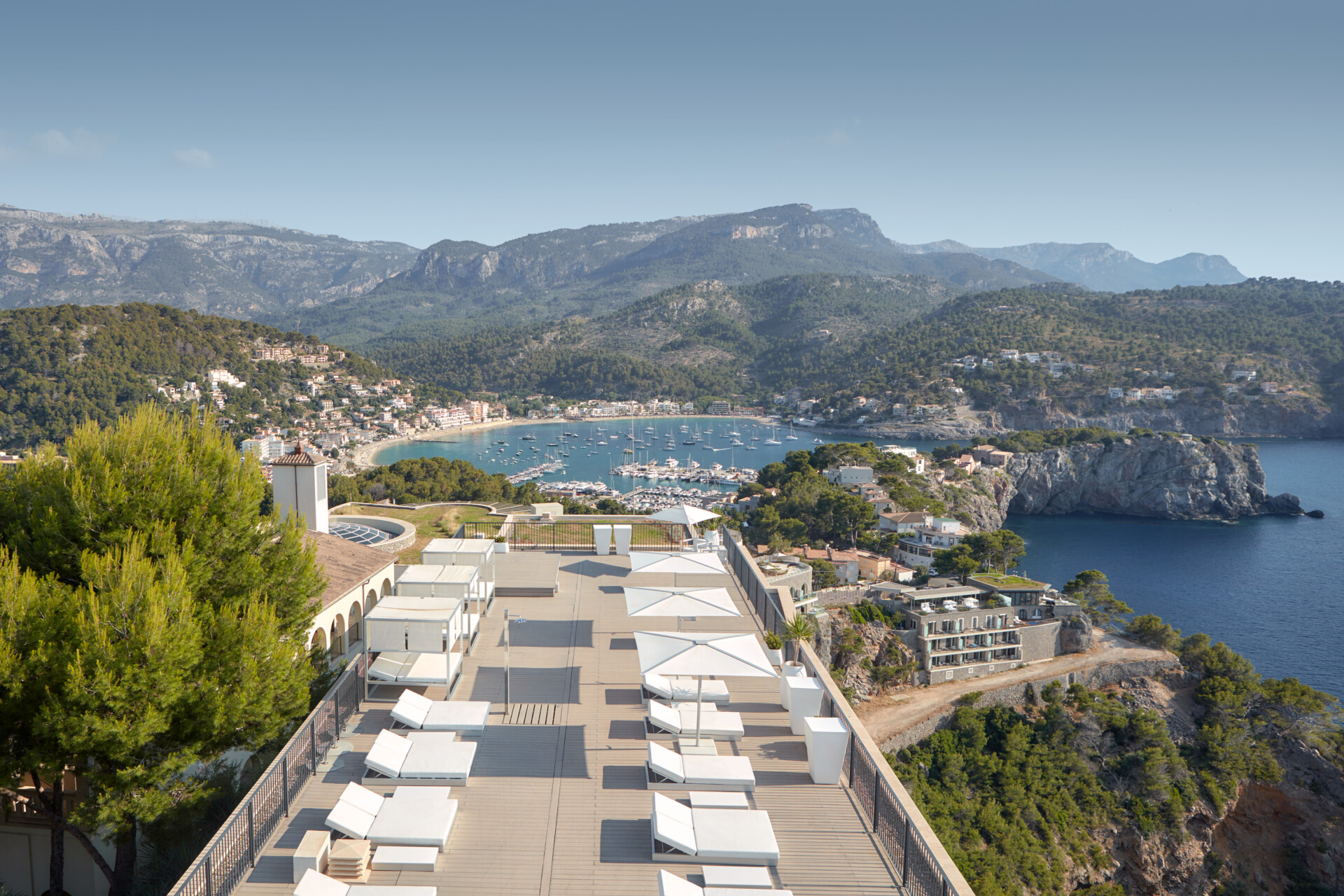 Panoramablick von einer erhöhten Terrasse mit Liegestühlen, die auf das azurblaue Wasser einer malerischen Bucht blicken, umgeben von grünen Hügeln und einer felsigen Küstenlinie mit Gebäuden und vereinzelten Booten im Wasser.