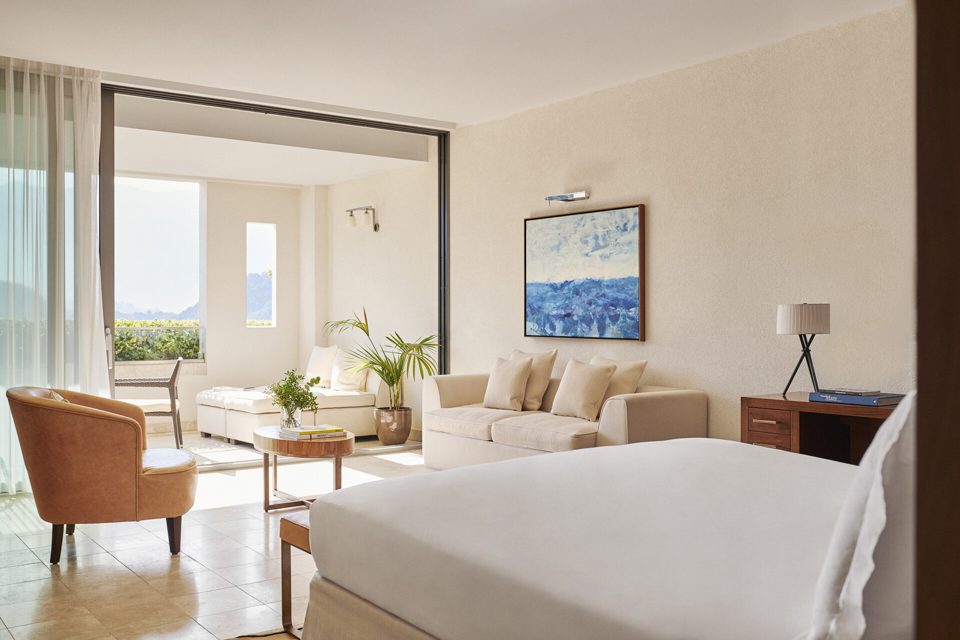 Ein helles und einladendes Hotelzimmer mit geschmackvoller Einrichtung. Im Vordergrund sieht man ein breites, gemütlich aussehendes Bett. Dahinter befindet sich eine Sitzecke mit einem beigen Sofa und einem passenden Sessel. Ein Couchtisch mit einer Glasplatte und frischen Pflanzen steht zwischen den Sitzmöbeln. An der Wand hängt ein abstraktes Gemälde mit blauen und weißen Farbtönen. Auf der rechten Seite ist ein Holzschreibtisch mit einer modernen Tischlampe platziert. Große Fenstertüren führen auf einen Balkon, der einen Blick auf eine malerische Landschaft mit Bergen im Hintergrund freigibt. Die Szene wird durch natürliches Sonnenlicht, das durch die Fenster hereinstrahlt, erhellt.