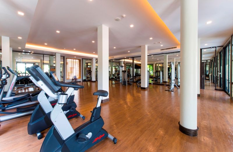 Fitnessraum im Layana Resort & Spa Ein heller und geräumiger Fitnessraum mit Holzboden, weißen Säulen und modernen Trainingsgeräten wie Laufbändern und Fahrrad-Ergometern. Die Beleuchtung ist warm und einladend, und große Spiegel bedecken die Wände, wodurch der Raum größer wirkt.