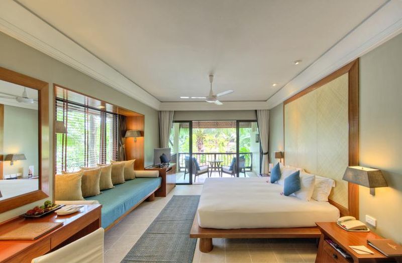Garden Pavilion im Layana Resort & Spa Ein helles und geräumiges Hotelzimmer mit großem Fenster und Blick auf eine Terrasse. Das Zimmer verfügt über ein großes Doppelbett mit weißen Laken und blauen Akzentkissen, eine lange Bank mit vielen Kissen, einen Arbeitstisch mit Stuhl, seitliche Nachttische mit Lampen und einen Deckenventilator. Der Boden ist mit einem dunklen Teppich ausgelegt und die natürliche Beleuchtung unterstreicht die warme und einladende Atmosphäre.