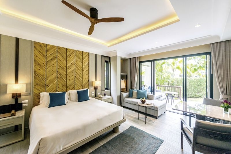 Grand Garden Pavilion im Layana Resort & Spa Ein helles und stilvoll eingerichtetes Hotelzimmer mit einem großen Doppelbett und blauen Akzentkissen, einer Sitzgruppe mit einem Sofa und einem Couchtisch im Vordergrund. Im Raum sorgen Holzelemente und indirekte Beleuchtung für eine warme Atmosphäre. Große Fenstertüren führen auf einen Balkon mit Blick ins Grüne.