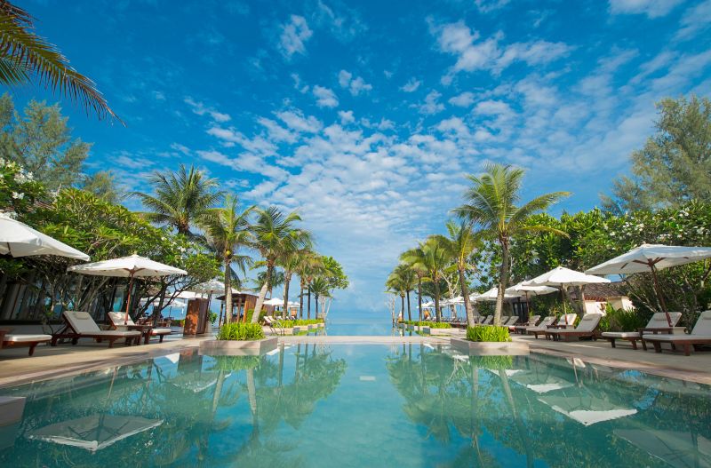 Infinitypool im Layana Resort & Spa Ein luxuriöser Swimmingpool mit klarem blauem Wasser im Vordergrund, umgeben von stilvollen Liegen und weißen Sonnenschirmen. Reihen von hohen Palmen säumen den Pool, der nahtlos in den sichtbaren Ozean übergeht. Der Himmel ist weit und blau mit malerischen weißen Wolken. Es strahlt ein Gefühl von Ruhe und exotischer Entspannung aus.