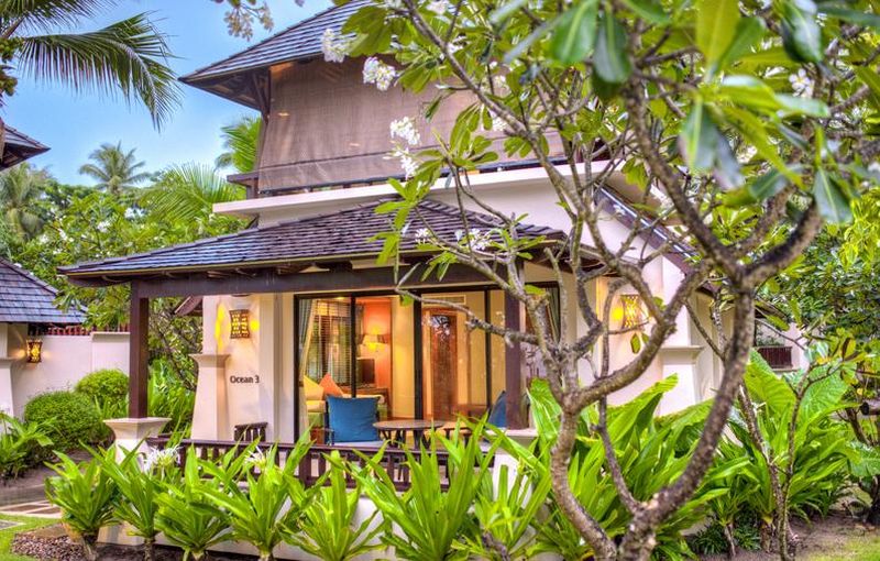 Ocean Deluxe Villa im Layana Resort & Spa Ein charmantes Ferienhaus im asiatischen Stil, versteckt hinter üppigem Grün mit blühenden Pflanzen und Palmen. Das Haus verfügt über ein klassisches Giebeldach und eine einladende Veranda mit einer blauen Sitzgelegenheit. Es ist Abendstimmung mit sanft eingeschaltetem Licht im Inneren, was eine warme und gemütliche Atmosphäre schafft.