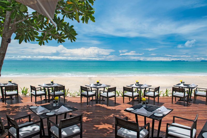 Restaurant Tides im Layana Resort & Spa Einladendes Strandrestaurant im Freien mit mehreren gedeckten Tischen auf einer Holzterrasse, mit makellosem Blick auf einen ruhigen blauen Ozean und einen hellen Sandstrand unter einem klaren Himmel. Lushes Grün grenzt an die Terrasse und sorgt für ein natürliches Ambiente.