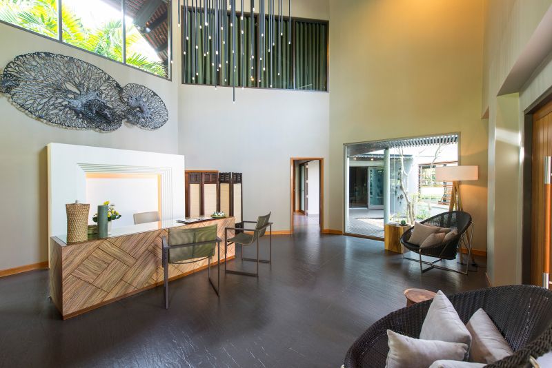 Rezeptionsbereich im Layana Resort & Spa Ein modern gestaltetes Hotel-Lobby-Interieur mit hohen Decken und dunklem Holzboden. An der Vorderseite befindet sich ein stilvoller Empfangstisch, begleitet von minimalistischen Sitzgelegenheiten und dekorativen Pflanzen. Über dem Empfangstisch hängt ein großes, kunstvolles Wandobjekt, das an ein Korallenriff erinnert. Große Fenster sorgen für natürliches Licht und Blick auf einen Außenbereich.