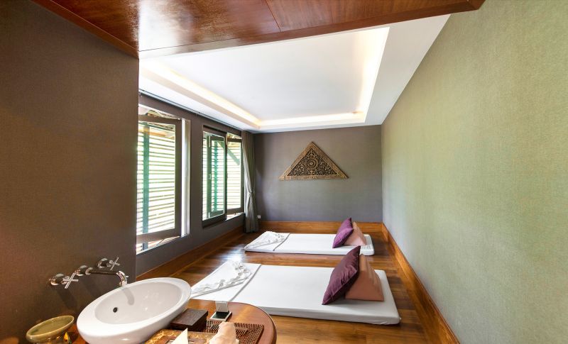 Spa Bereich im Layana Resort & Spa Ein harmonisch gestaltetes, modernes Badezimmer mit integrierter Schlafgelegenheit. Das Zimmer zeigt eine weiße freistehende Badewanne, ein Waschbecken mit Spiegel und eine erhöhte Plattform mit einer Matratze und Kissen. Natürliches Licht fällt durch Jalousien an den Fenstern. Warme Holzelemente an Decke und Boden sowie eine dekorative Wandkunst vervollständigen das Ambiente.