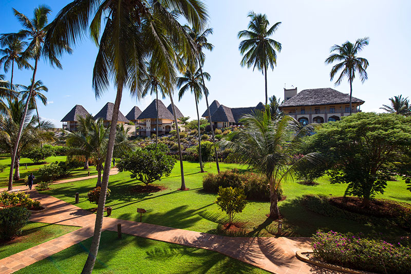Ein tropisches Resort umgeben von saftig grünen Rasenflächen und hohen Palmen unter einem klaren blauen Himmel. Im Hintergrund sind strohgedeckte Dachspitzen und das Hauptgebäude des Resorts sichtbar.