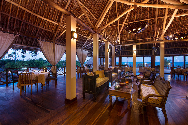 Ein elegantes Restaurant mit traditioneller Holzarchitektur und Strohdach. Mehrere Tische sind für das Essen vorbereitet, und gemütliche Sitzbereiche bieten einen Blick auf das Meer und die Palmen im Hintergrund. Die Beleuchtung ist warm und einladend, was eine entspannende Atmosphäre schafft.