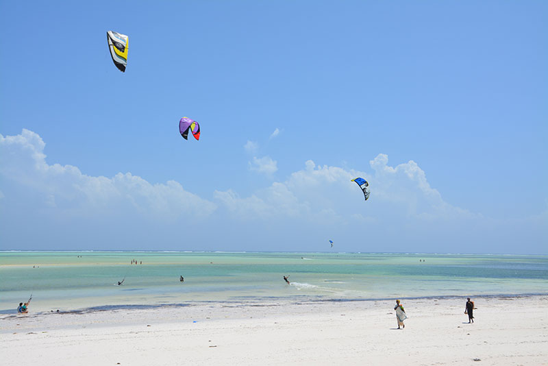 Bild eines sonnigen Strandtages mit mehreren Kitesurfern, die farbenfrohe Kites über dem seichten türkisfarbenen Meerwasser steuern. Personen schlendern am weißen Sandstrand, während im Hintergrund ein klarer blauer Himmel vorherrscht.