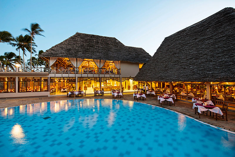 Abendansicht eines luxuriösen Resorts mit Strohdächern, das eine offene Gaststätte und belegte Tische an einem großen, einladenden Swimmingpool zeigt. Palmen säumen das Bild und schaffen eine tropische Stimmung.