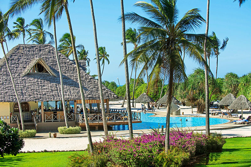 Ein tropisches Resort mit Strohdächern, einer großen Poolanlage und umgeben von Palmen unter einem klaren blauen Himmel. Im Vordergrund blühen violette Blumen und sattgrünes Gras.