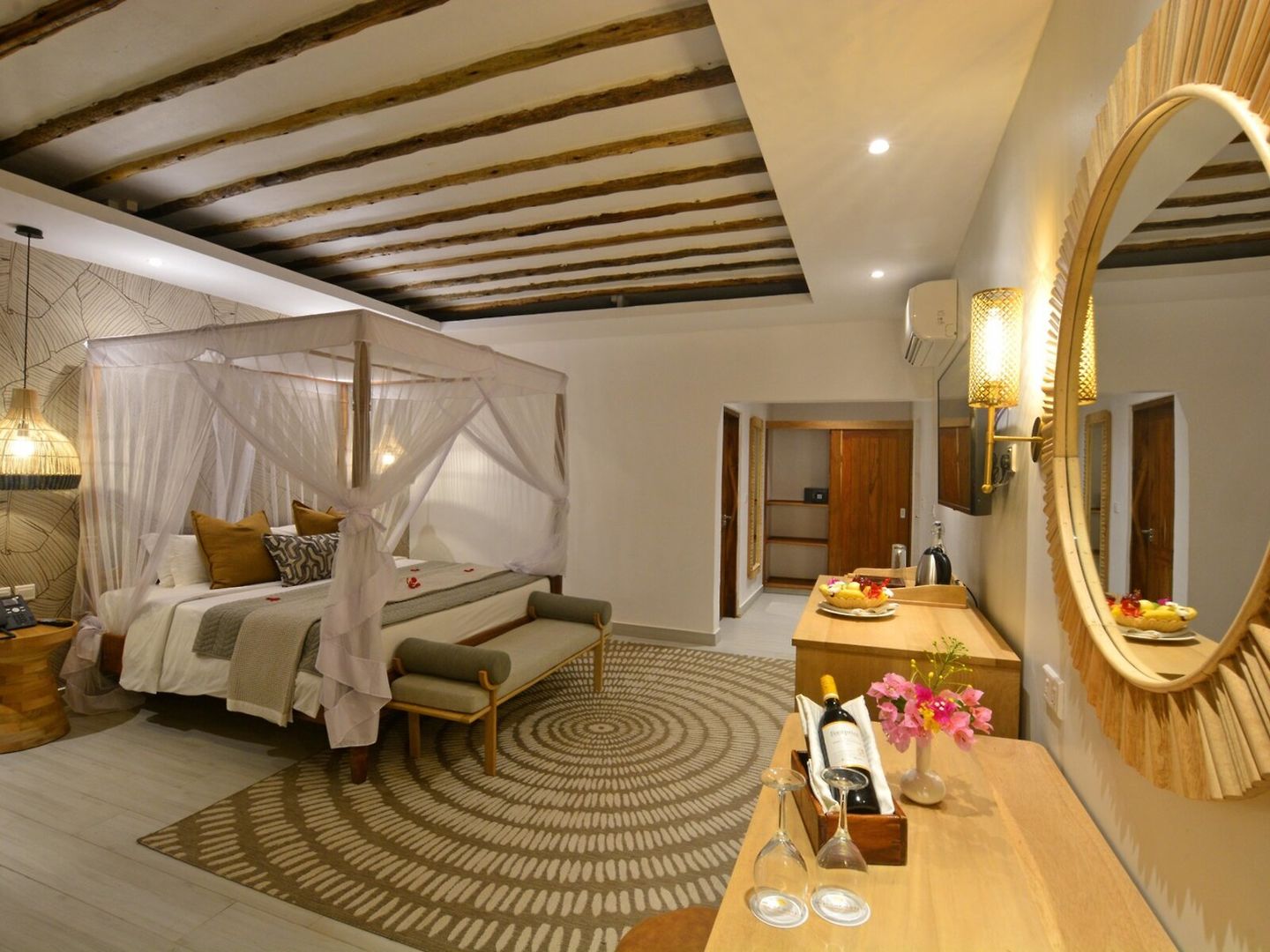 Ein geräumiges und stilvoll eingerichtetes Schlafzimmer mit einem großen Himmelbett, umgeben von einem zarten, weißen Moskitonetz. Über dem Bett befinden sich rustikale Holzbalken, die sich harmonisch in das moderne Design einfügen. Eine elegante Sitzbank am Fußende des Bettes, dekorative Kissen und eine weiche Tagesdecke runden den Komfort ab. Im Vordergrund ist ein hölzerner Tisch sichtbar, darauf eine Flasche Wein mit zwei Gläsern und ein Teller mit frischem Obst. An der Wand hängt ein großer, runder Spiegel, der das warme Licht einer stylischen Wandlampe reflektiert. Im Hintergrund führen zwei Türen zu weiteren Räumen.