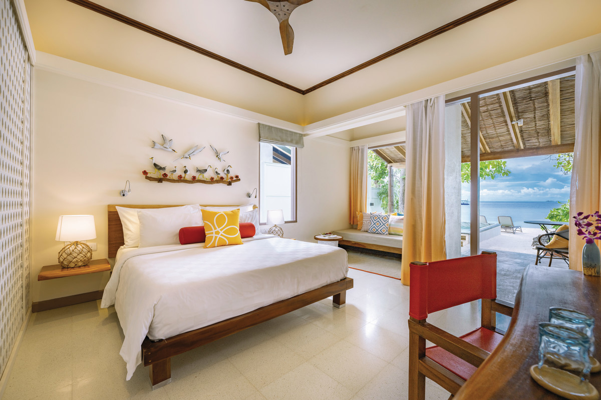 Deluxe Beach Villa Heller und einladender Strandhotelzimmer mit einem gemütlichen Doppelbett, eingerichtet mit farbenfrohen Kissen und einer Kunstinstallation mit Vögeln über dem Bett. Große Fenstertüren öffnen sich zu einer Terrasse mit Aussicht auf einen tropischen Strand.