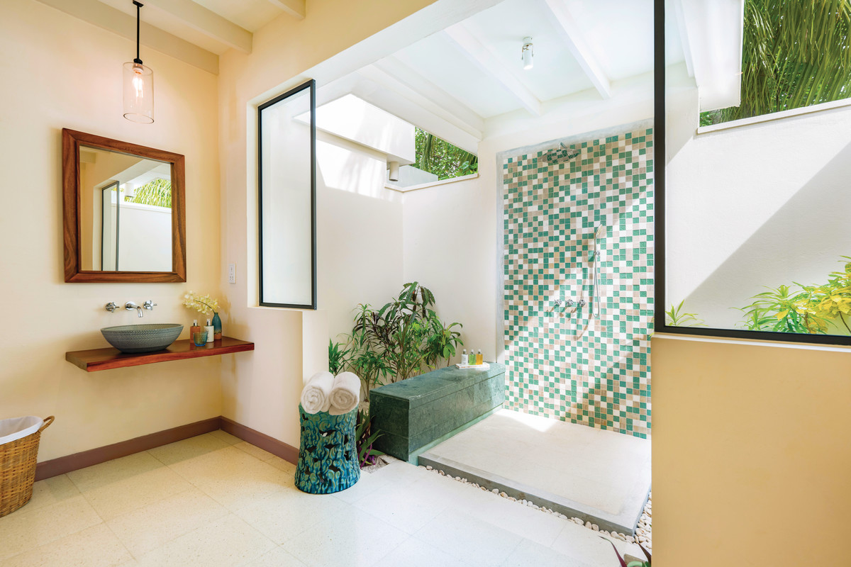 Badezimmer in einer Deluxe Beach Villa Ein helles, modernes Badezimmer mit einer Duschkabine, deren Wandfliesen grünlich und beige Mosaikmuster aufweisen. Eine freistehende Wanne befindet sich gegenüber einer Fensterfront, die viel natürliches Licht hereinlässt und einen Blick auf üppiges Grün außerhalb des Raums ermöglicht. Unter einem großen Spiegel an der Wand steht ein Waschtisch mit einer dunklen Schüssel und Armaturen, daneben ein kleiner Abfallkorb und eine stilvolle Vase mit frischen Handtüchern.