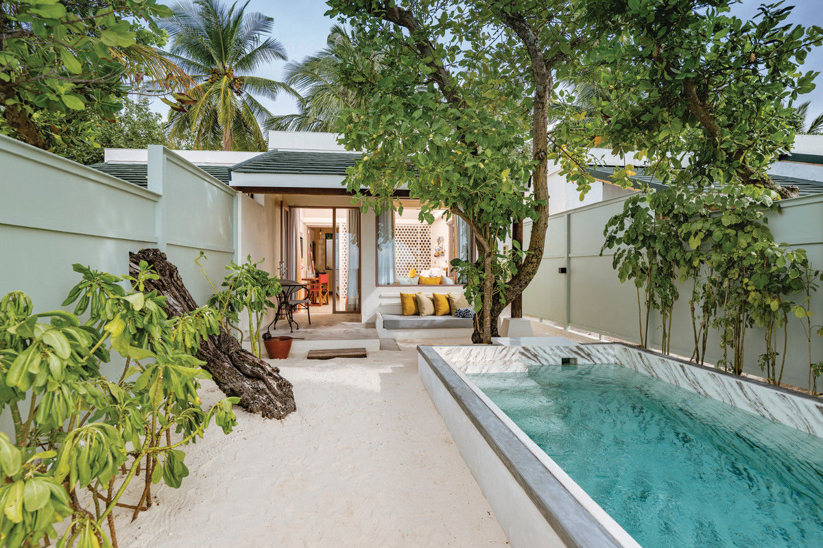 Deluxe Beach Villa mit Pool Ein idyllischer Rückzugsort im Freien mit einem privaten Swimmingpool an einem sandigen Strand. Der Pool ist von einer niedrigen Mauer eingefasst, neben Bäumen und tropischen Pflanzen. Ein modernes Feriendomizil mit offener Tür lädt im Hintergrund ein, während draußen eine Sitzgelegenheit mit Kissen für Entspannung sorgt.