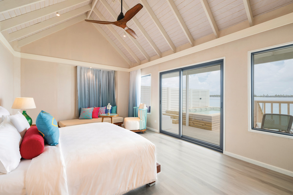 Lagoon Villa mit Pool Ein helles und luftiges Strandhotelzimmer mit einem Kingsize-Bett, farbenfrohen Kissen, Holzdeckenventilator und bodentiefen Fenstern, die auf einen Balkon mit Blick auf ruhiges Wasser führen.