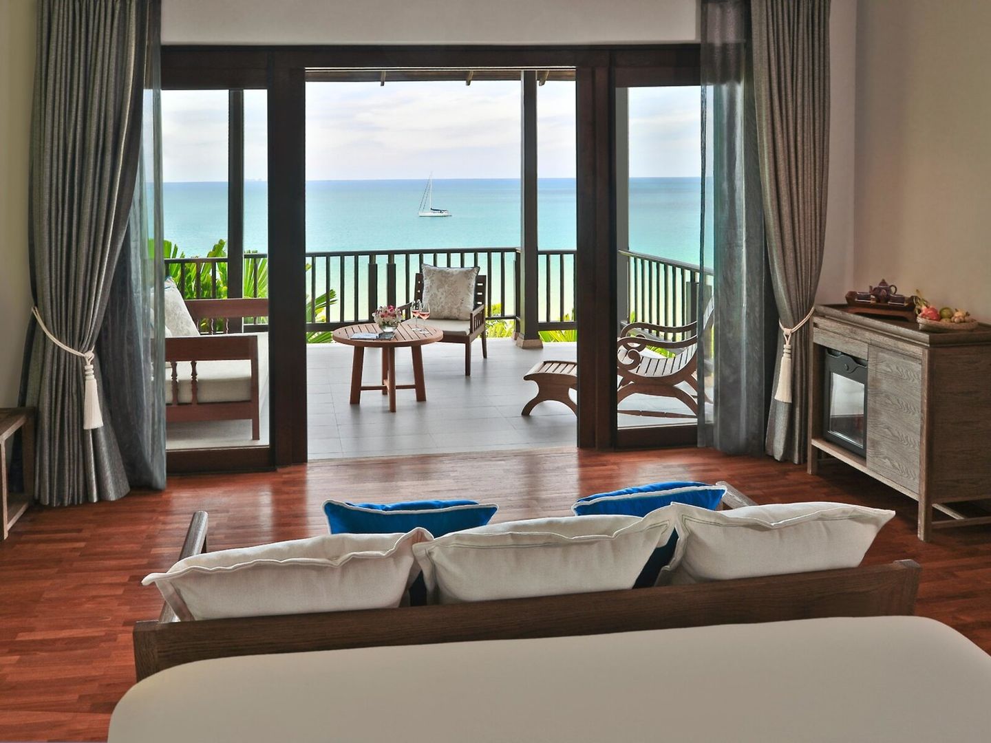 Ausblick von einem Wohnbeispiel Ein luxuriöses Schlafzimmer mit Blick auf das Meer durch geöffnete Glasschiebetüren, die zu einem Balkon mit Sitzgelegenheiten führen. Die Inneneinrichtung umfasst ein großes Bett im Vordergrund mit weißen Kissen, Holzböden, schwere Vorhänge und elegante Möbel. Im Hintergrund ist eine Yacht auf dem ruhigen, blauen Meer zu sehen.