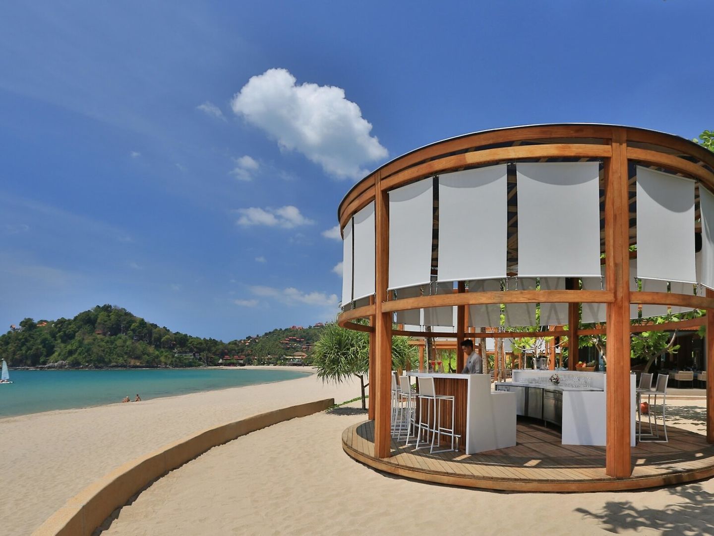 Bar Bild eines runden Strand-Bars mit Holzstruktur und großen Glasfenstern auf Sand, mit Blick auf eine ruhige Bucht, umgeben von grüner Vegetation unter einem blauen Himmel mit einzelnen Wolken.