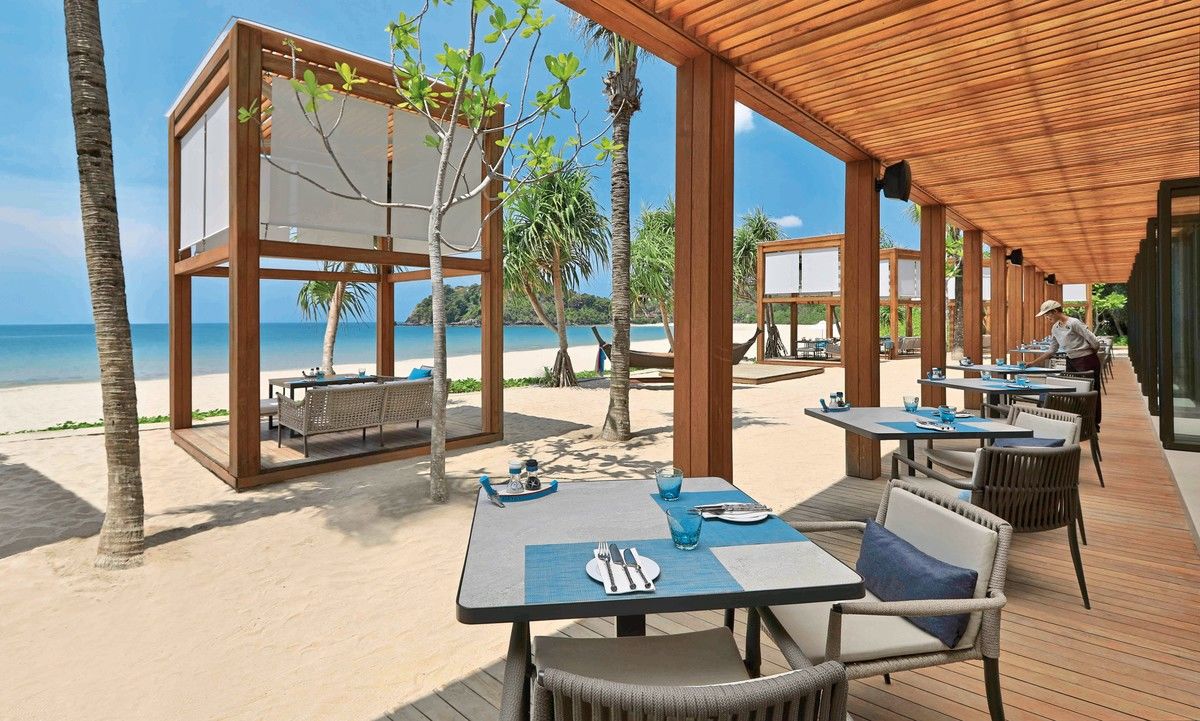 Ray Talay Bar & Restaurant Ein elegantes Restaurant im Freien mit Blick auf einen ruhigen Strand. Die Einrichtung umfasst moderne Tische und Stühle auf einer hölzernen Terrasse, geschützt durch ein Dach aus Holzbalken. Palmen säumen den weißen Sandstrand, der sich bis zum klaren Meereswasser erstreckt. Eine entspannte Atmosphäre durch das natürliche Licht und den luftigen Raum wird betont.