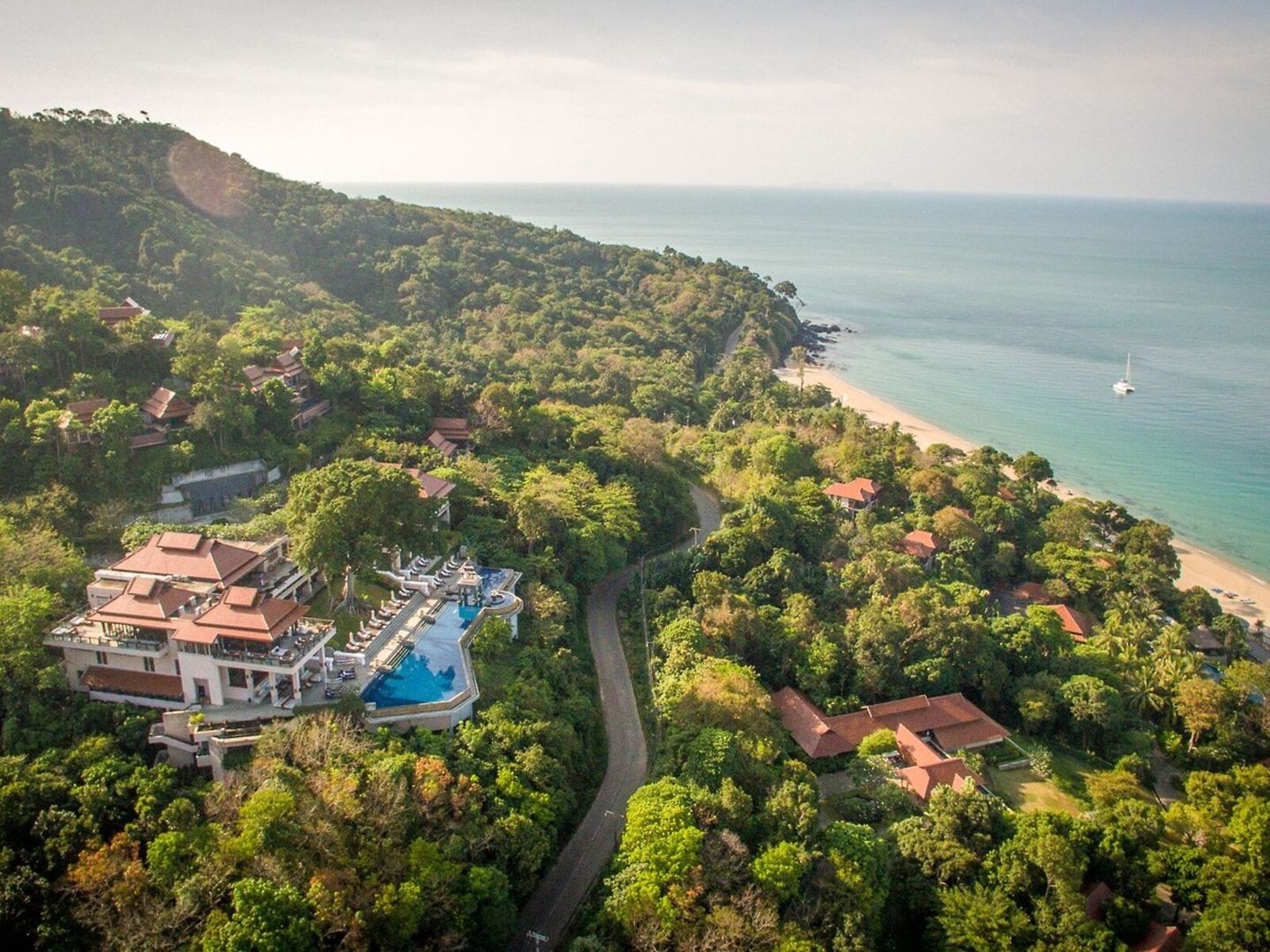 Blick auf das Pimalai Resort & Spa Luftaufnahme eines luxuriösen Resorts am Hang mit üppiger Vegetation, mehreren Pools, und angrenzendem Strand am klaren blauen Meer mit einem Segelboot auf dem Wasser.