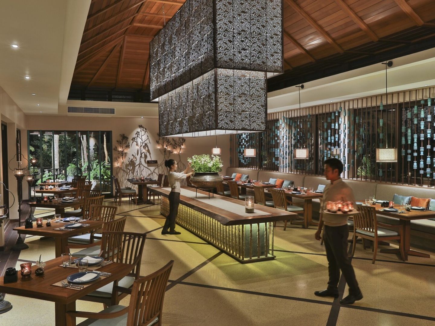 Restaurant Ein stilvolles Restaurant mit modernem Interieur. Im Vordergrund Holztische mit elegant gedeckten Plätzen. Bedienungen bewegen sich zwischen den Tafeln, eine trägt Teller. Die hohe Decke zeigt sichtbare Holzbalken und große Hängeleuchten. Im Hintergrund sind durch großflächige Fenster Bäume zu erkennen, was für ein natürliches Ambiente sorgt.