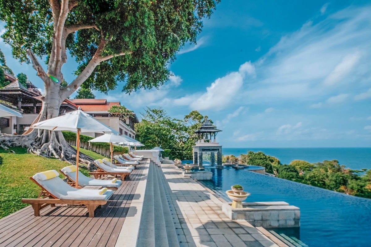 Sonnenliegen am Pool Ein luxuriöser Außenbereich eines Resorts mit einem Infinity-Pool, der über den Horizont zu schweben scheint. Sonnenliegen mit gelben Handtüchern sind unter weißen Schirmen auf einer Holzterrasse aufgereiht. Ein markanter Baum bietet Schatten und das Meer ist im Hintergrund sichtbar, was ein Gefühl von Ruhe und Exklusivität vermittelt.