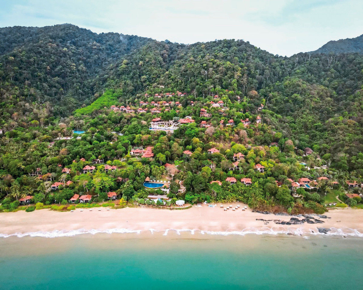 Blick auf das Pimalai Reosrt & Spa und den Strand Luftaufnahme eines tropischen Strandresorts mit üppiger Vegetation und dichtem Wald im Hintergrund. Rote Dachziegel heben sich von den verschiedenen Grünschattierungen der Bäume ab, während ein sanfter Strand die Szene abschließt.