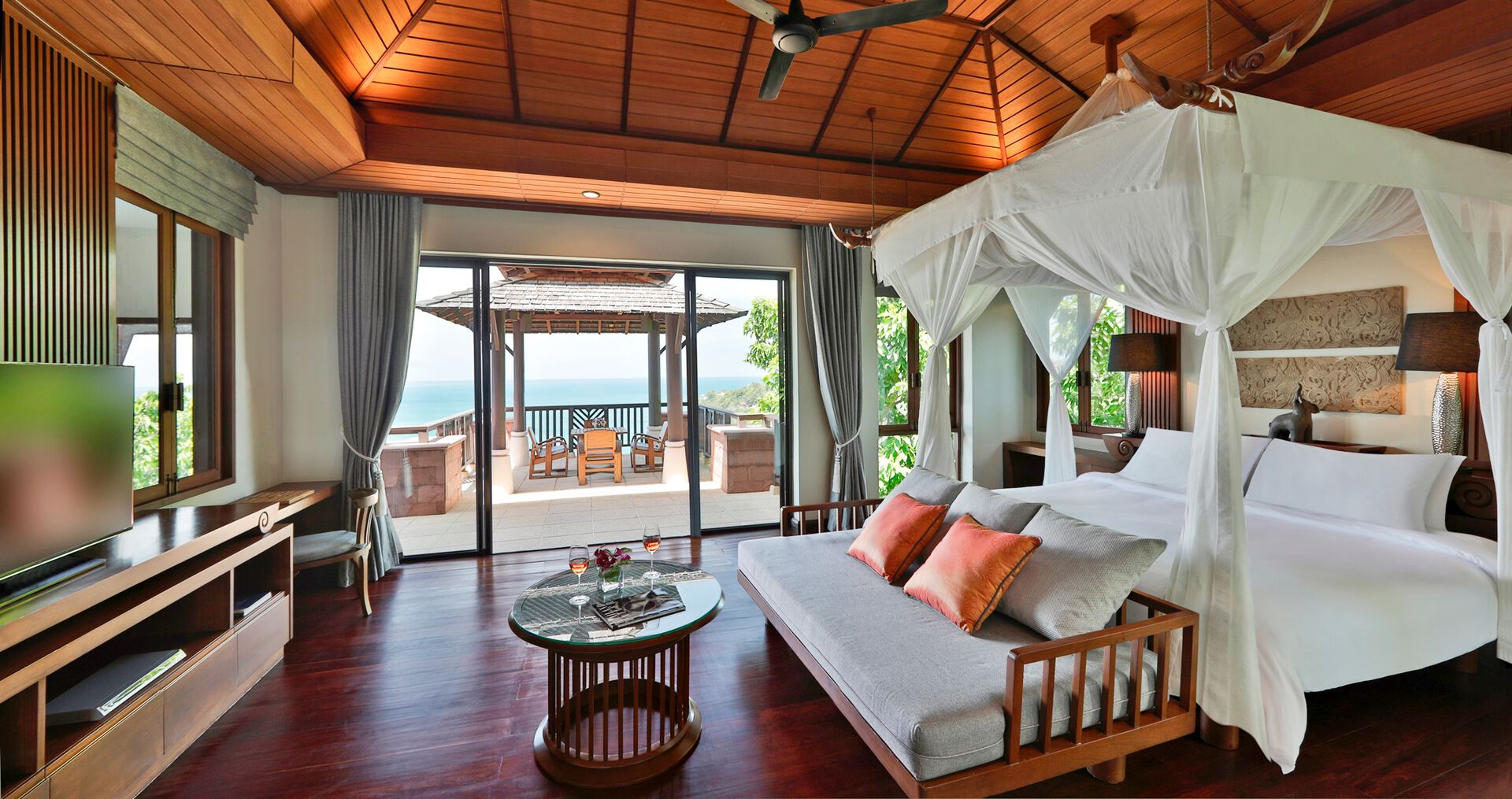 Wohnbeispiel Ein luxuriöses Schlafzimmer in einem Ferienresort mit Holzboden und -decke, einem großen Himmelbett und einer Lounge-Ecke. Große Fenster bieten einen beeindruckenden Blick auf das Meer und führen zu einer privaten Terrasse mit Sitzgelegenheiten im Freien.
