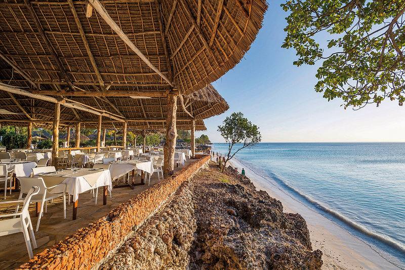 Strandrestaurant mit weiß eingedeckten Tischen unter einem großen Strohdach, gelegen an einem ruhigen blauen Meer neben einem felsigen Ufer, umgeben von tropischer Vegetation.